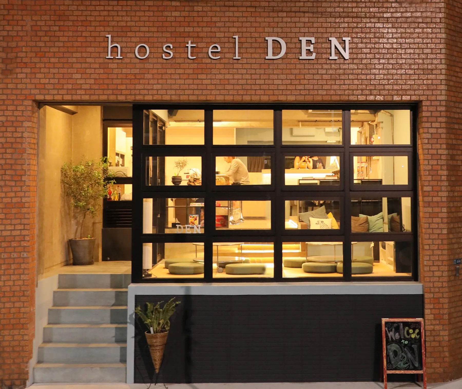 夜のhostel DENの外観。赤レンガの壁と、黒い枠の大きな窓の奥に明かりのついたラウンジ